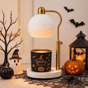 May include: A gold-toned candle warmer lamp with a white dome shade featuring cut-out patterns. The base is white with the text CASAMATA printed on it. A black candle jar sits on the base, decorated with orange pumpkin and bat graphics and the text Spooky Season.