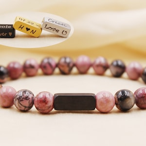 May include: A pink and black beaded bracelet with a black metal bar charm. The charm is engraved with the words "Nicole Forever" and "07.22 H ♥ N".