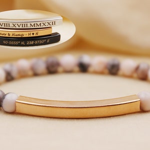 Peut inclure: Un bracelet en or avec un charme en forme de barre gravé de chiffres romains, les mots "Forever & Always", un symbole de cœur et les initiales "H & N". Le bracelet est composé de perles avec un motif marbré rose et gris.