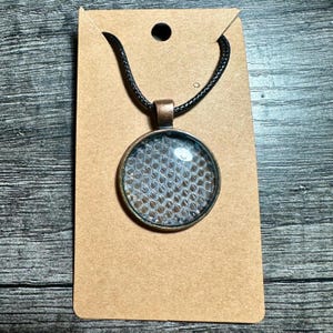 Peut inclure: Un collier pendentif rond avec un cadre de couleur bronze et un cordon noir. Le pendentif présente un dôme en verre transparent sur un motif texturé de couleur claire. Le collier est présenté sur une carte marron.