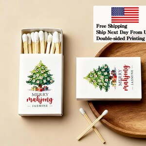 May include: White matchbox with a watercolor Christmas tree design, the text "Merry Mahjong - Jasmine" in red. The box is open, revealing matches with white tips. A second box and matches are also visible.