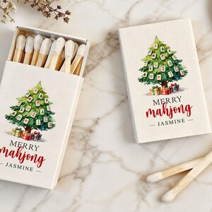 May include: Two boxes of matches with a Christmas tree design. The tree is decorated with mahjong tiles and presents. The text "MERRY mahjong - JASMINE" is printed in red. One box is open, revealing the matchsticks.
