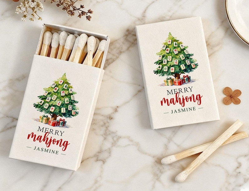 May include: Two boxes of matches with a Christmas tree design. The tree is decorated with mahjong tiles and presents. The text "MERRY mahjong - JASMINE" is printed in red. One box is open, revealing the matchsticks.