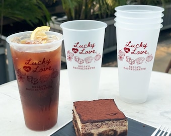 Puede incluir: Un vaso de plástico transparente lleno de té helado con una rodaja de limón, junto a tres vasos vacíos. Todos los vasos tienen el texto "Lucky in Love" y "Bella's Bachelorette" impreso en rojo. Una porción de tiramisú está en un plato negro.
