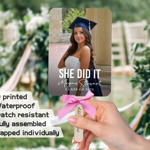 Pu&ograve; includere: Un ventaglio di laurea rosa e bianco con una foto di una donna con cappello e toga. Il ventaglio dice "She Did It" con il nome del laureato e l'anno di laurea. Il ventaglio &egrave; tenuto da una mano.