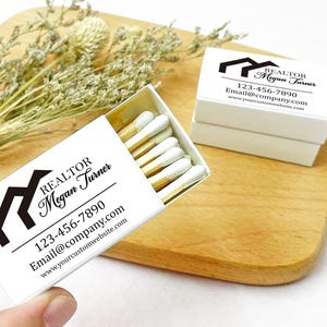 May include: White matchbox with a black house icon and the text "REALTOR Megan Turner" with contact information. The matchbox is on a wooden surface.