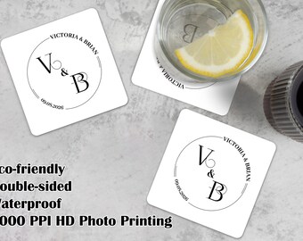 May include: Three square white coasters featuring a black circular monogram design with the names Victoria & Brian and the date 09.08.2026. One coaster is placed under a glass containing a lemon slice, while two others are displayed separately.
