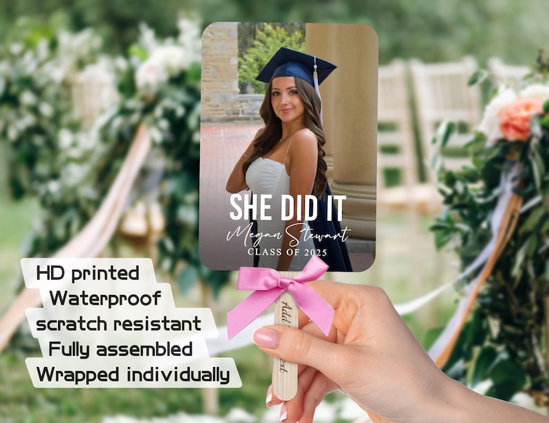 Pu&ograve; includere: Un ventaglio di laurea rosa e bianco con una foto di una donna con cappello e toga. Il ventaglio dice "She Did It" e include il nome del laureato e l'anno di laurea. Il ventaglio &egrave; tenuto da una mano.