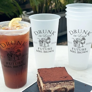 May include: Three clear plastic cups with the text "DRUNK in LOVE EST. 2026 FUTURE MRS. BROWN" printed on them. The cups are filled with a clear liquid. A slice of lemon is in one of the cups.