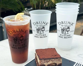 May include: Three clear plastic cups with the text "DRUNK in LOVE EST. 2026 FUTURE MRS. BROWN" printed on them. The cups are filled with a clear liquid. A slice of lemon is in one of the cups.