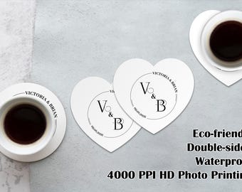 May include: Two white heart-shaped coasters and one round white coaster, each featuring a black circular design with the initials V & B, the names Victoria & Brian, and the date 09.09.2026. Two white ceramic mugs filled with black coffee are present.