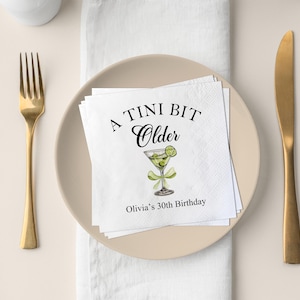 Könnte beinhalten: Eine weiße Serviette mit schwarzem Text, der "A Tini Bit Older" lautet, mit einer Martini-Glas-Illustration und dem Text "Olivia's 30th Birthday".