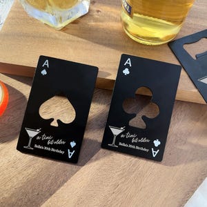 May include: Two black card-shaped bottle openers with a spade and a clover design. Each opener has a cut-out for a bottle cap and the text "a tiny bit older Bella's 30th Birthday".