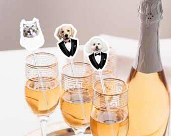 May include: Champagne flutes filled with a golden liquid, adorned with decorative stirrers featuring a cat and two dogs in formal attire. A champagne bottle is in the background. The flutes have gold rims.