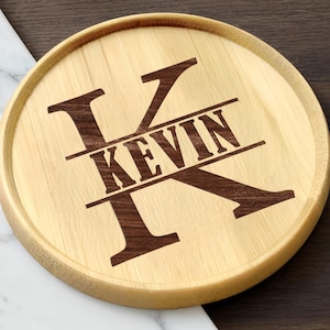 Puede incluir: Un posavasos redondo de madera clara. Presenta una gran letra "K" marrón oscuro con el nombre "KEVIN" grabado en una fuente a juego en el centro. El posavasos tiene un borde elevado.