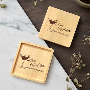 May include: Two square wooden coasters with a martini glass design and the text "a tini bit older, Julia's 40th Birthday". The coasters are light brown and have a square shape with a slightly recessed center.