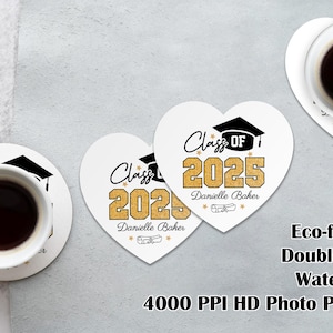 May include: Two white heart-shaped coasters with black text that reads "Class of 2025" and "Danielle Baker". The coasters are decorated with gold glitter and a black graduation cap. The coasters are on a light gray surface.