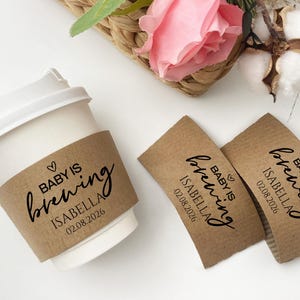 May include: A white paper coffee cup with a brown sleeve that reads "Baby is brewing" with the name Isabella and the date 02.08.2026. Two additional brown sleeves with the same text are also present. The image has a baby shower theme.