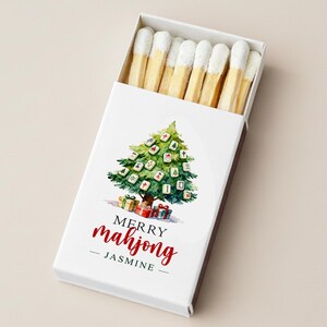 May include: A white matchbox with a watercolor Christmas tree design made of mahjong tiles. The text "MERRY mahjong - JASMINE" is written in red script. The box contains matches with white tips.