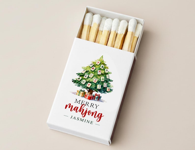 May include: A white matchbox with a watercolor Christmas tree design made of mahjong tiles. The text "MERRY mahjong - JASMINE" is written in red script. The box contains matches with white tips.