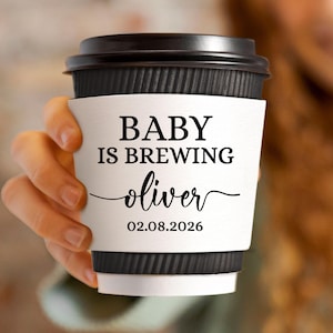 May include: A black takeaway coffee cup with a white sleeve. The sleeve features the text "BABY IS BREWING" above the name "oliver" and the date "02.08.2026" in black script. The cup is held by a person.