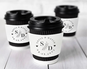May include: Three black disposable coffee cups with lids, each featuring a white sleeve. The sleeves display the names "SOFIA & DAVID", initials "S D", a sprig design, and the date "09.08.2025". The cups are on a white wooden surface.