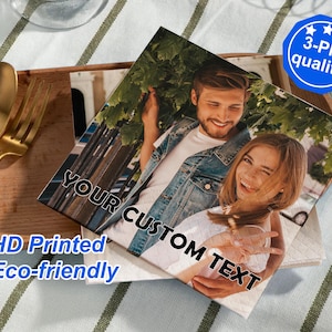 Può includere: Tovaglioli di carta quadrati con una foto stampata di una coppia sorridente. I tovaglioli hanno il testo "YOUR CUSTOM TEXT", "HD Printed" e "Eco-friendly". Un distintivo blu indica "3-Ply quality". I tovaglioli sono su un vassoio di legno con posate dorate.