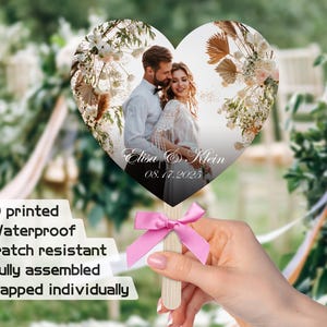 May include: A heart-shaped wedding fan with a photo of a couple in front of a floral arch. The fan is printed with the couple's names, "Elisa & Klein", and the date "08.17.2025".