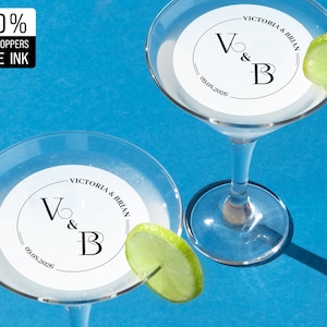 May include: Two white martini glasses with edible toppers featuring a black circle with the initials "V & B" and the date "09.05.2026" printed in black. The text "VICTORIA & BRIAN" is printed above the circle. A lime wedge is placed on the rim of each glass.