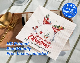 May include: A white paper napkin featuring a watercolor illustration of two martini glasses filled with red liquid, a small snowman, and red ornaments. The text reads Cheers to Christmas THE THOMPSON FAMILY in red and black script.