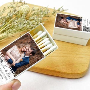 Puede incluir: Caja de cerillas blanca con una foto de una pareja en la parte delantera. El texto "Save Our Date" está impreso en la caja, junto con los nombres de la pareja, la fecha "August 25th, 2026" y la ubicación "San Diego, CA". También se imprime un código QR en la caja.