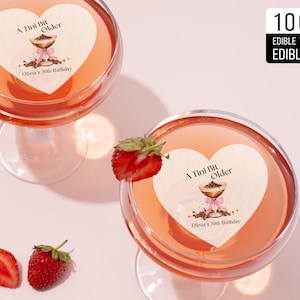 May include: Two heart-shaped edible cake toppers with the text "A Tini Bit Older" and "Olivia's 30th Birthday". The toppers are on top of martini glasses filled with a pink liquid. The glasses are on a white surface with strawberries.
