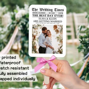 May include: A wedding fan with a newspaper design featuring a photo of a couple and the text "The Wedding Times", "Denver, Colorado", "08.17.2025 4:00 PM", "The Best Day Ever!", "Elisa & Klein", "Are Getting Married", "Today is the day they will say "I Do"". The fan is made of sturdy cardstock and has a pink ribbon attached to the handle.
