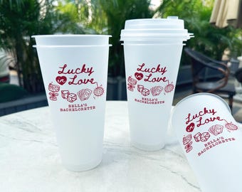 Puede incluir: Tres vasos de plástico blanco con el texto "Lucky in Love" y "Bella's Bachelorette" impreso en rojo. Los vasos están sobre una superficie blanca.