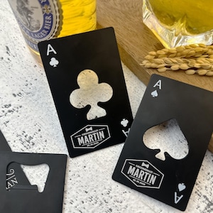Puede incluir: Abrebotellas de metal negro con forma de cartas de juego, As de Picas y As de Tréboles. El trébol tiene un recorte en forma de trébol, y la pica tiene un recorte en forma de pica. Cada carta tiene el nombre "MARTIN" y una fecha.