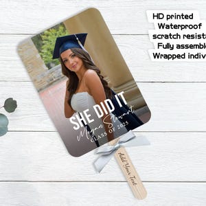 Pu&ograve; includere: Un ventaglio di laurea con una foto di una donna con cappello e toga. Il ventaglio ha il testo "She Did It" con il nome del laureato e l'anno della sua classe. Il ventaglio &egrave; bianco con un nastro argentato.