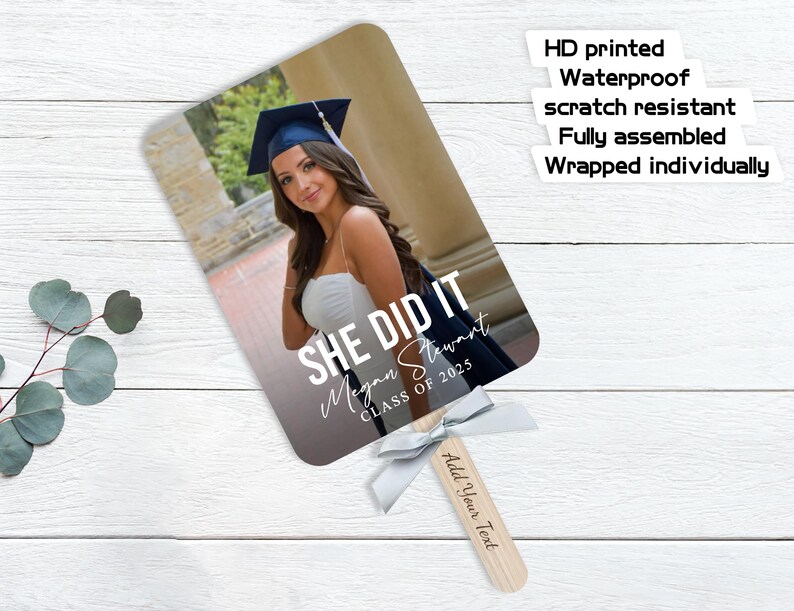 Pu&ograve; includere: Un ventaglio di laurea con una foto di una donna con cappello e toga. Il ventaglio ha il testo "She Did It" con il nome del laureato e l'anno della sua classe. Il ventaglio &egrave; bianco con un nastro argentato.