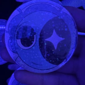 May include: A circular pin-back button featuring a stylized graphic of a crescent moon face with a sleepy expression. The design is rendered in shades of blue and black with a holographic star pattern overlay that reflects light under blue illumination.