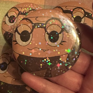 May include: A circular pinback button featuring a tan-coloured cartoon character with large eyes, round glasses, and a small fang. The surface has a holographic finish with a scattered star-shaped glitter pattern that reflects various colours.