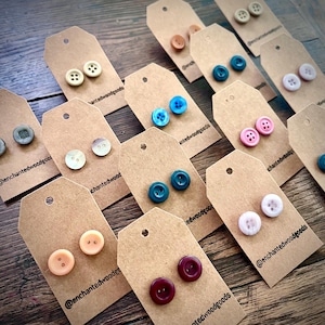 May include: Assortment of colorful button earrings displayed on brown card stock with the text "@enchantedwoodgoods" on each card. The buttons are various colors, including pink, blue, green, red, and beige.
