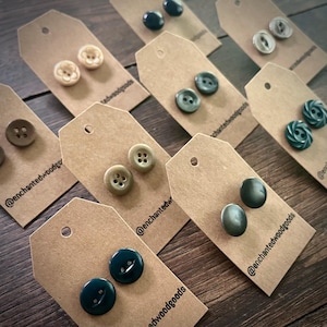 May include: Assortment of vintage buttons displayed on brown paper cards with the text "@enchantedwoodgoods" on each card. The buttons are in various colors and patterns, including green, brown, black, and white.