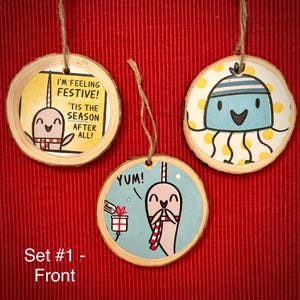 May include: Set of three wooden Christmas ornaments. Each ornament features a cartoon narwhal or jellyfish design with festive text. The ornaments are round with a natural wood edge and a twine hanger. The background is red.