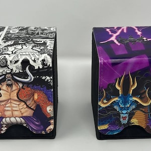 May include: Two black card deck boxes with different anime character designs. The box on the left features a black and white manga style image of a character with red and purple accents. The box on the right features a purple and black image of a dragon with gold accents.