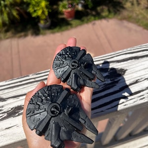 May include: Two black Millennium Falcon models are held in a hand. The models are detailed with circular and rectangular shapes, and have a textured surface. The background shows a wooden deck and greenery.