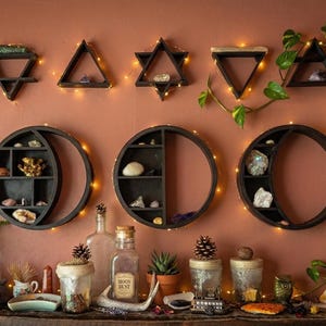 以下が含まれることがあります： 温かみのある壁に設置された、三角形、星、円など、さまざまな幾何学的な形状の暗い木製棚のコレクション。 棚にはクリスタルやその他の装飾品が置かれています。 フェアリーライトと緑が美しさを添えています。