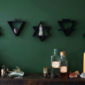 Könnte beinhalten: Eine dunkelgrüne Wand zeigt fünf schwarze geometrische Regale mit Kristallen. Darunter befindet sich ein Holzregal mit Pflanzen, Flaschen, einer Kerze und Büchern. Die Szene erinnert an ein mystisches oder spirituelles Thema mit Fokus auf natürliche Elemente und Dekoration.