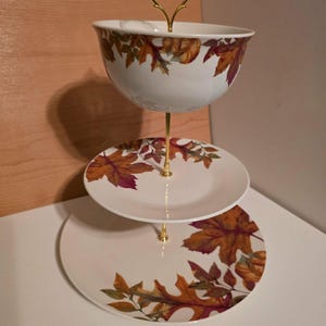 May include: A three-tier serving stand featuring a white bowl on top and two white plates below, all decorated with a colorful autumn leaf pattern in shades of orange, brown, and red. The stand has a gold-colored metal central rod and handle.