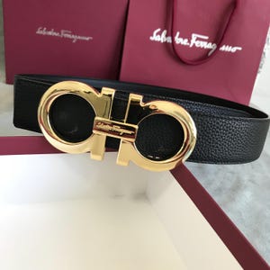 May include: A black textured leather belt featuring a polished gold-tone metal Gancini buckle. The buckle displays the Salvatore Ferragamo brand name engraved in the centre. The belt strap has a pebbled finish and is positioned horizontally.