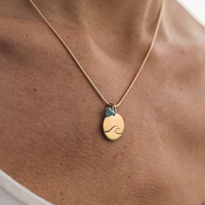 Op de afbeelding: Een gouden ketting met een kleine turquoise hanger en een ronde hanger met een golfontwerp.