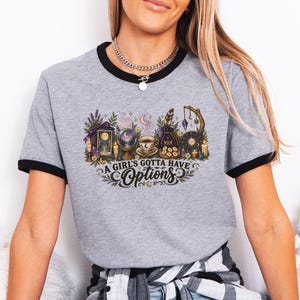 May include: A heather gray ringer t-shirt with black trim at the neck and sleeves. The front features a graphic print of mystical items including a crystal ball, tarot card, and coffee cup, with the text A GIRL'S GOTTA HAVE OPTIONS printed below.
